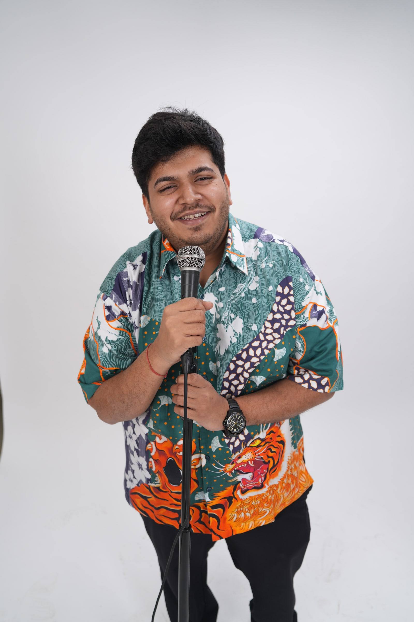 Kaustubh Agarwal smiling at the camera, holding a microphone on a stand, in a printed green shirt