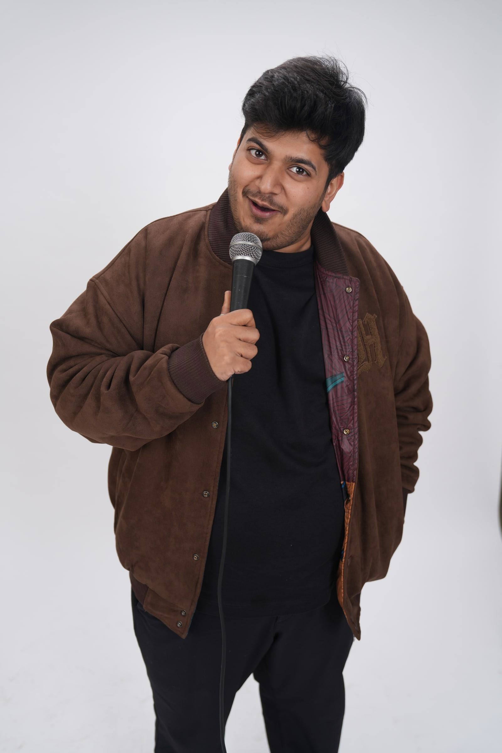 Kaustubh Agarwal in a brown jacket, holding a microphone and pulling a face at the camera
